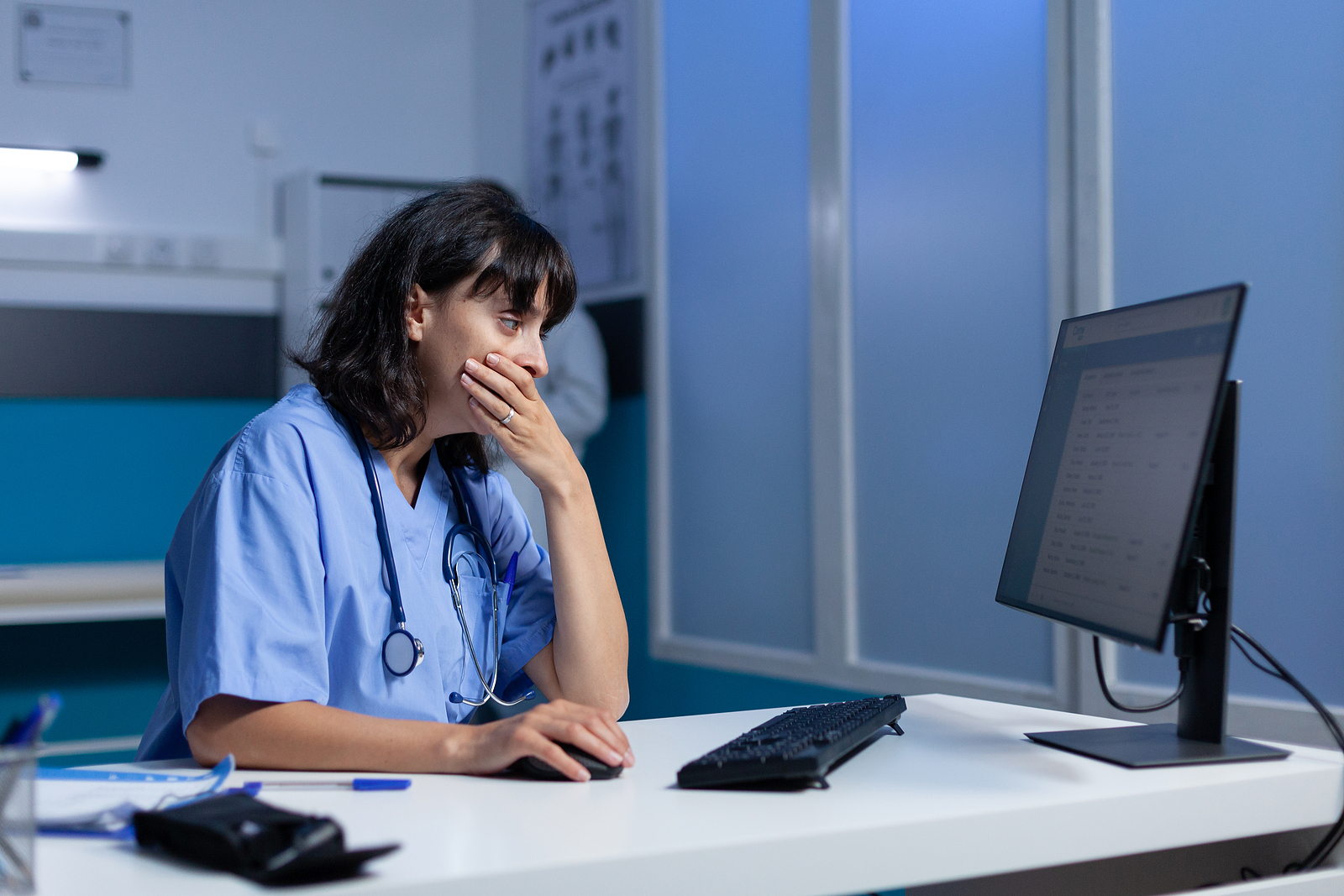 Tired nurse using computer on desk while falling asleep, working late. Medical assistant looking at monitor screen and feeling sleepy after overtime work at night in healthcare cabinet.